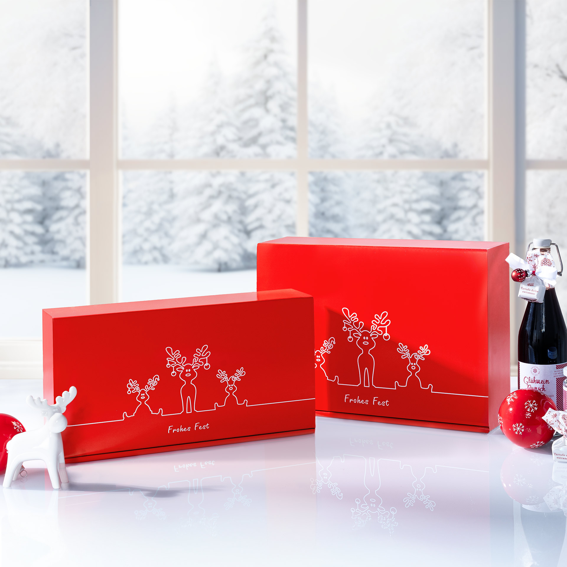 Präsentkarton - Rentier Trio - 2 x 0,75 l Rote Geschenkboxen mit Rentier Trio Design vor einem winterlichem Fenster.